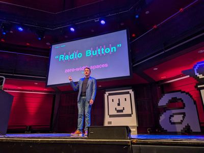 Leo Vogel on stage in a blazer and jeans, pointing at a large screen showing the words “Radio Button” split by zero-width spaces, beside a giant pixel-art retro computer.